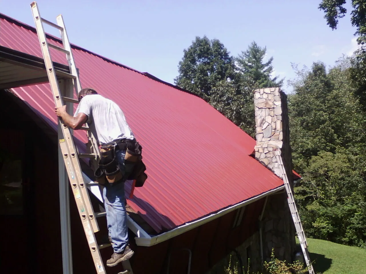 Roofer on a ladder beside a red metal roof and stone chimney — Soffit & Fascia Repair by Roofing Brothers in Westwood